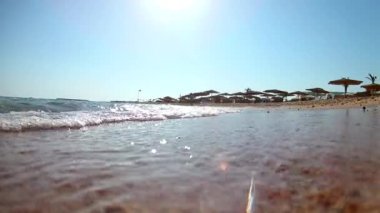 Açık güneşli bir günde sazdan plaj şemsiyeleri ile kumlu plajda sörf ışık dalgaları sörf, Kızıldeniz, aşağıdan görünümü, geniş açı. Yavaş çekim Standart hareket.