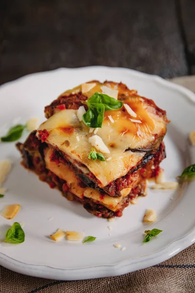 Tradizionale Parmigiana di melanzane: melanzane al forno - Italia, cucina siciliana.Melanzane al forno con formaggio, pomodori e spezie su un piatto bianco. Un piatto di melanzane è su un tavolo di legno — Foto Stock