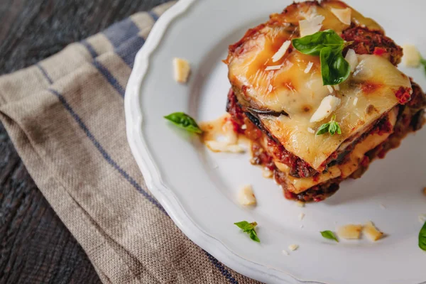 トラディショナルパルミジャーナ・ディ・メランザン:焼きナス - イタリア、シチリアのいとこ。チーズ、トマト、スパイスを白い皿に焼いたナス。ナスの皿は木製のテーブルの上にある — ストック写真