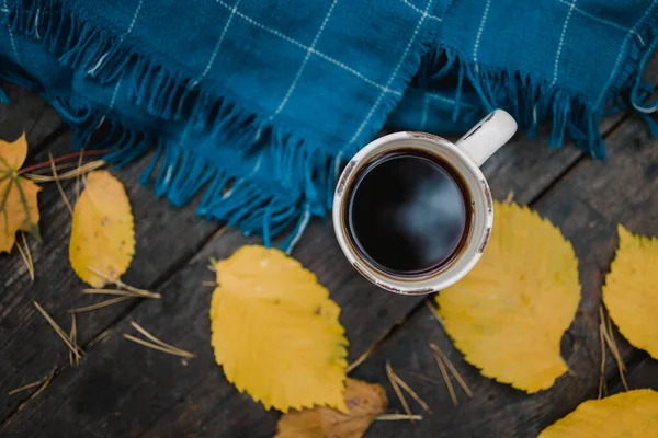 En una vieja mesa de madera en el parque de otoño hay una taza de té y café. Una bufanda a cuadros azul cálido se dispersa con hojas amarillas y conos de pino. Vista superior, borrosa. Otoño cálido estado de ánimo oscuro, espacio de copia — Foto de Stock