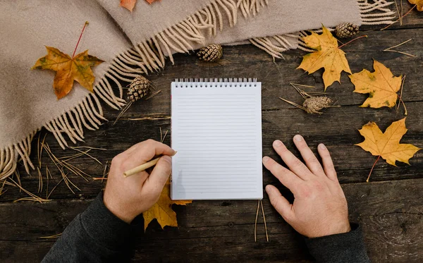 Menschenhände mit einer Tasse Kaffee und Schal am Holztisch mit Notizbuch und Stift und Herbstblättern. Menschenhände mit einer Tasse Kaffee und einem Schal am Holztisch — Stockfoto