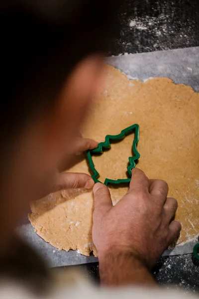 Male hands cut out gingerbread cookie in the form of a snowflake, Christmas tree, a man from raw dough on parchment baking paper on a dark background. View from above. save space — Stock Photo, Image