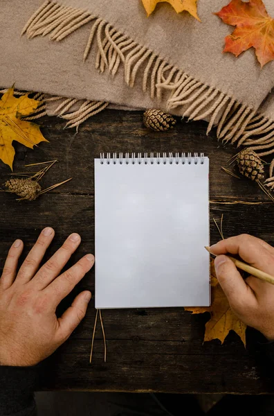 Menschenhände mit einer Tasse Kaffee und Schal am Holztisch mit Notizbuch und Stift und Herbstblättern. Menschenhände mit einer Tasse Kaffee und einem Schal am Holztisch. — Stockfoto