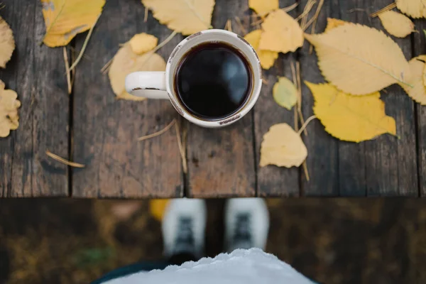 En una vieja mesa de madera en el parque de otoño hay una taza con té y café, hojas amarillas dispersas y conos de pino. Vista superior, las piernas visibles en desenfoque. Otoño cálido estado de ánimo oscuro, espacio de copia — Foto de Stock