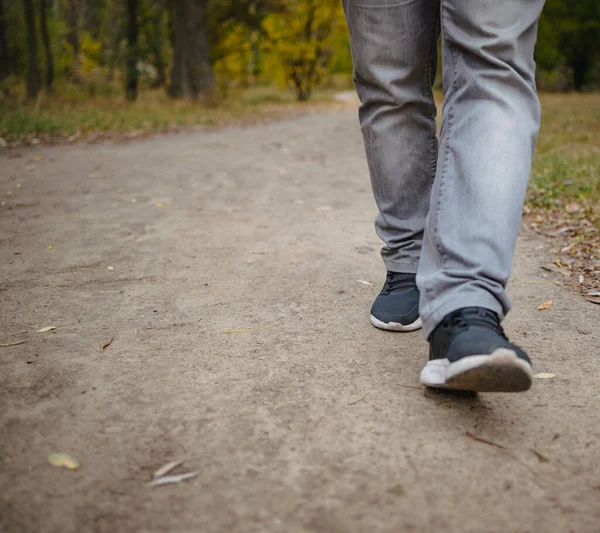Mannelijke benen in jeans en sneakers lopen in de verte op de grond bedekt met gevallen gele bladeren. Herfst aankomst concept, close-up, kopieerruimte. — Stockfoto