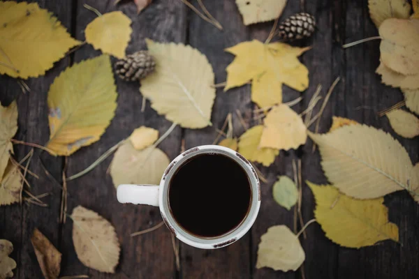Su un vecchio tavolo di legno nel parco autunnale c'è una tazza di tè e caffè, foglie gialle sparse e pigne. Vista dall'alto, sfocata. Autunno caldo umore scuro, soft focus . — Foto Stock