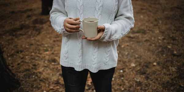 Jeune femme dans un pull blanc tient une tasse de thé avec du café sur un fond de feuilles d'automne jaunes. Ambiance automnale chaude, mise au point douce. Espace de copie — Photo