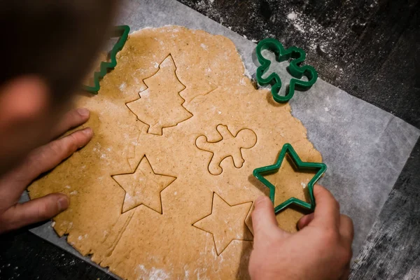 male hands cut out gingerbread cookie in the form of a snowflake, Christmas tree, a man from raw dough on parchment baking paper on a dark background. View from above. save space