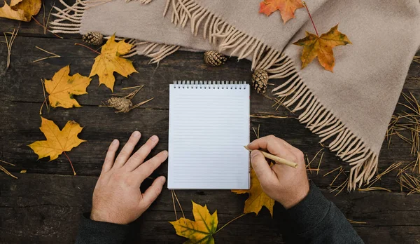 Manos humanas con una taza de café y bufanda en la mesa de madera con cuaderno y bolígrafo y hojas de otoño. Manos humanas con una taza de café y bufanda en la mesa de madera — Foto de Stock