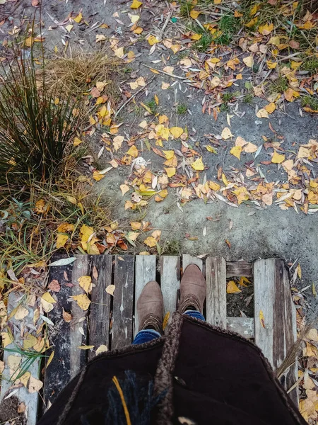 Pemandangan atas kaki wanita dengan sepatu bot coklat di latar belakang daun musim gugur kuning dan platform kayu. Salin ruang . — Stok Foto