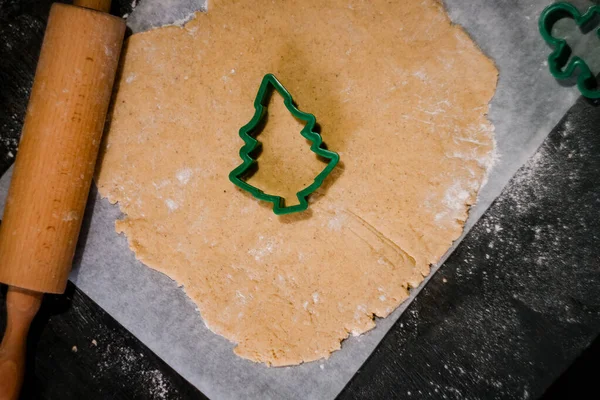 Corten la forma de la galleta de jengibre en forma del árbol de Navidad de la masa cruda sobre el papel de hornear pergamino sobre el fondo oscuro. Vista desde arriba. ahorrar espacio — Foto de Stock