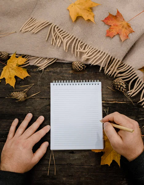 Manos humanas con una taza de café y bufanda en la mesa de madera con cuaderno y bolígrafo y hojas de otoño. Manos humanas con una taza de café y bufanda en la mesa de madera — Foto de Stock