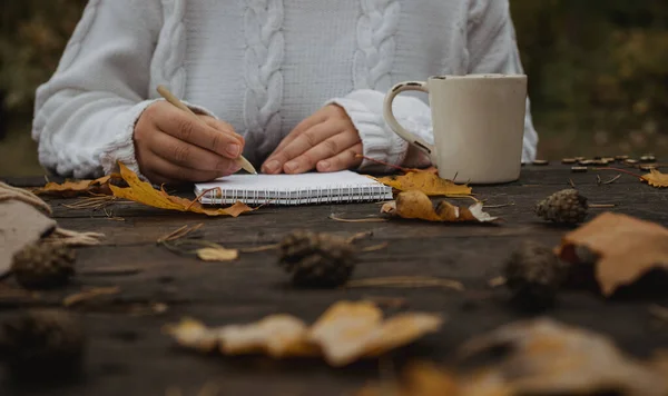 Human hands with a cup of coffee and scarf at wooden table with notebook and pen and autumn leaves. Human hands with a cup of coffee and scarf at wooden table