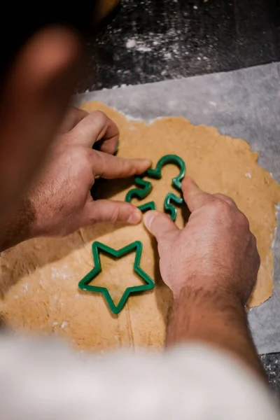 Male hands cut out gingerbread cookie in the form of a snowflake, Christmas tree, a man from raw dough on parchment baking paper on a dark background. View from above. save space — Stock Photo, Image