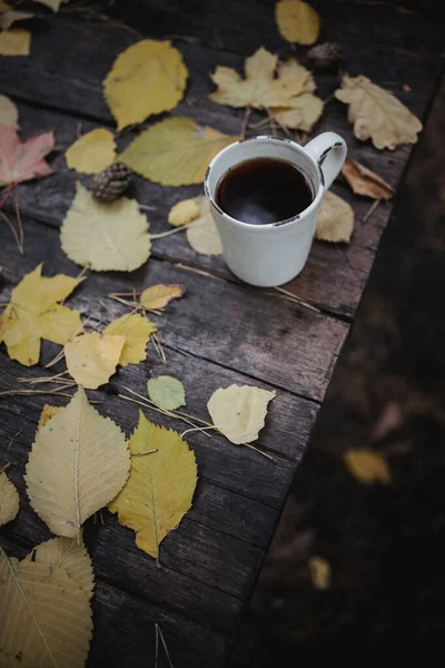 En una vieja mesa de madera en el parque de otoño hay una taza con té y café, hojas amarillas dispersas y conos de pino. Vista superior, borrosa. Otoño cálido humor oscuro, enfoque suave . — Foto de Stock