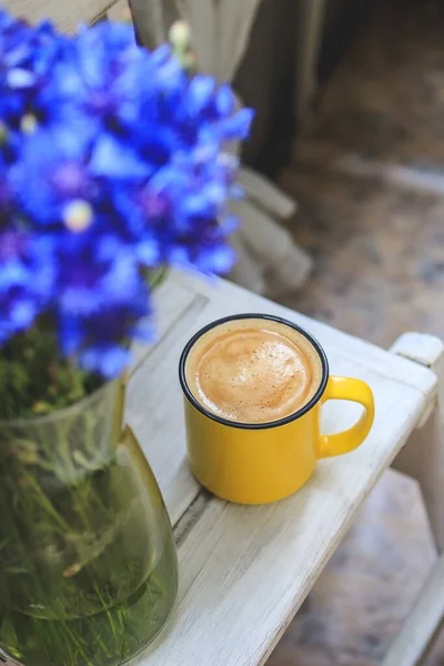 En el balcón hay una mesa de madera blanca con una taza de café amarillo y un jarrón con acianos. —  Fotos de Stock