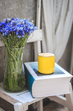 Balkonda beyaz ahşap bir masa, kalın bir kitap, sarı bir fincan kahve ve mısır çiçekli bir vazo var..