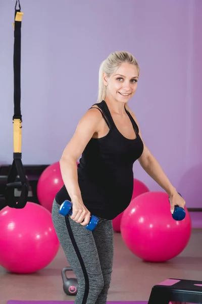 Mujer embarazada de pie en el gimnasio, sosteniendo sus pesas en 9 meses con hermosa vista y haciendo ejercicio sobre hombros y brazos — Foto de Stock