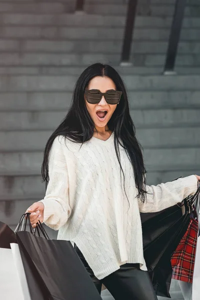 Hermosa chica en gafas de sol con bolsas de papel caminando fuera del centro comercial — Foto de Stock