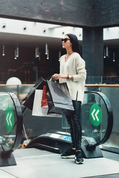 Menina bonita com sacos de papel na escada rolante no supermercado — Fotografia de Stock