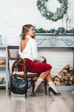 bir kırmızı etek ve beyaz kazak retro tekli süslenmiş Noel duvar arka plan üzerinde oturan güzel genç kadın. Erotik bacaklar beyaz çorap ve siyah cila yüksek heells