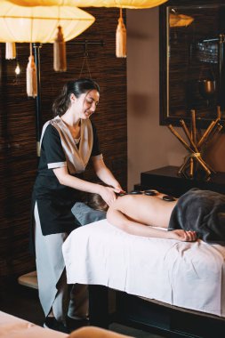 Uzman bir istemciye sıcak taş masajı yapma. Masaj masasında döşeme panchakarma yordam genç çekici kız. kabine modern spa rahatlatıcı, vakit geçirmek güzel bir kadın. Yumuşak sarı ışık.