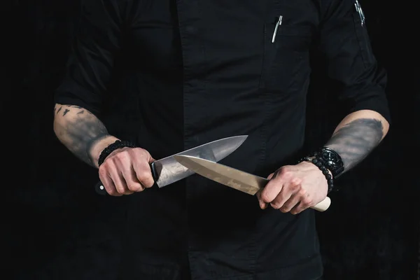 Genç çekici adam Şef dövmeler siyah üniforma elinde iki bıçak tutar. siyah arka plan