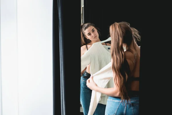 Young beautiful woman shopping, in a fitting room in fashion mall, trying on a white sweater — 스톡 사진