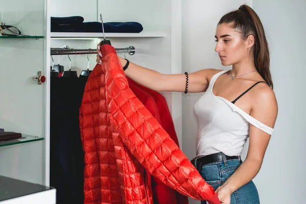 Young woman in a blu jeance white t-shirt trying on red down coat in a luxury boutique — Stock Photo, Image