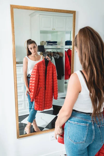 Young woman in a blu jeance white t-shirt trying on red down coat in a luxury boutique — Stock Photo, Image
