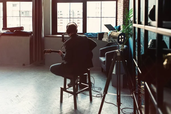 Un chanteur assis sur un tabouret dans un casque avec une guitare enregistrant une piste dans un home studio — Photo