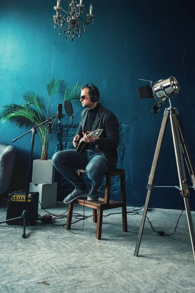Чоловік співак сидить на табуреті в навушниках з гітарою, записуючи трек в домашній студії — стокове фото