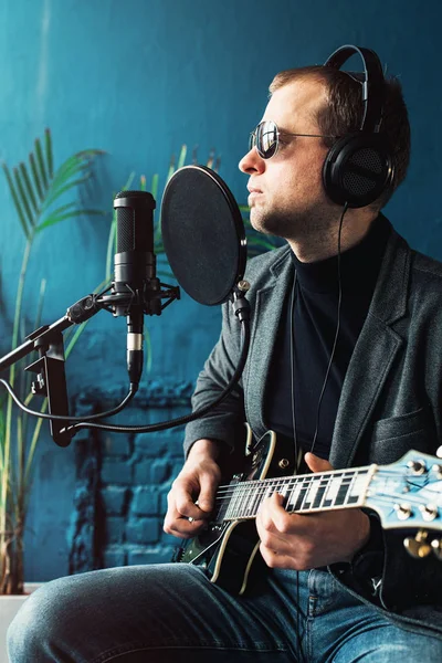Close up of a man singer sitting on a stool in a headphones with a guitar recording a track in a home studio
