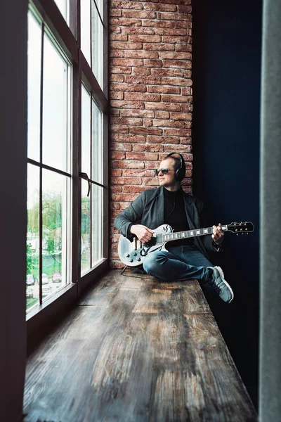 Чоловік співак сидить на підвіконні в навушниках з гітарою, записуючи трек в домашній студії — стокове фото
