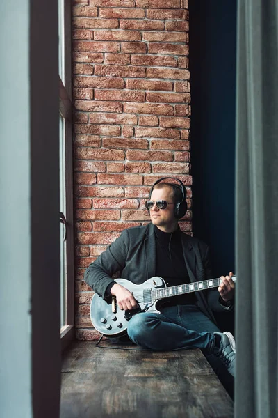 Чоловік співак сидить на підвіконні в навушниках з гітарою, записуючи трек в домашній студії — стокове фото