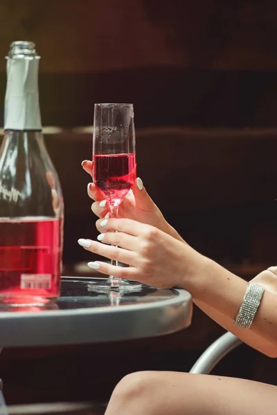 Jovem mão segurando um copo com champanhe rosa. Garrafa aberta em uma mesa — Fotografia de Stock