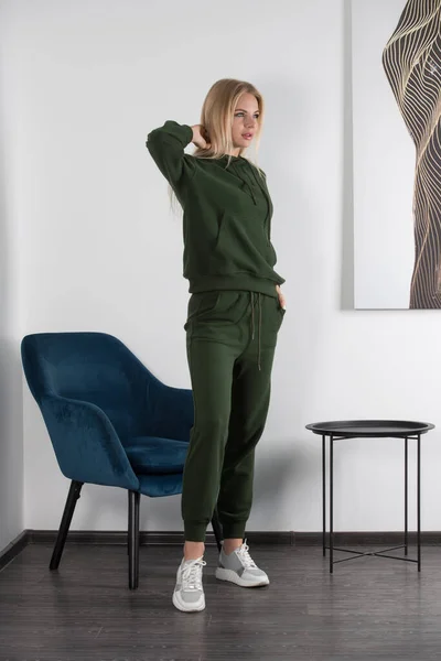 身穿绿色运动服的年轻貌美的金发女子在房间的白墙边摆姿势 迷人的女模特靠蓝色椅子摆姿势 健身女士 — 图库照片