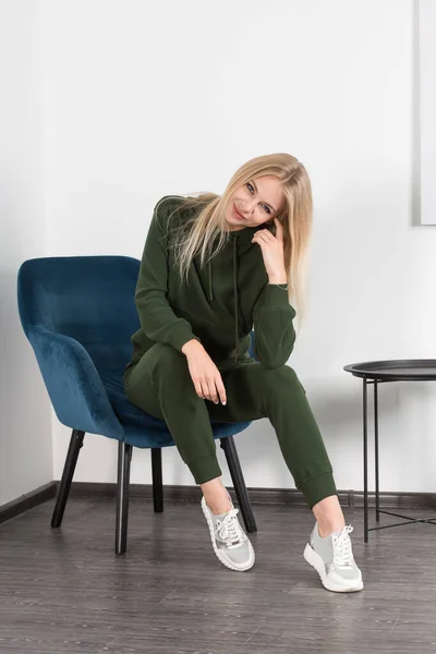 Mulher loira bonita e elegante em um traje verde posa perto de uma parede branca na sala. Modelo de menina atraente posando em uma cadeira azul. — Fotografia de Stock