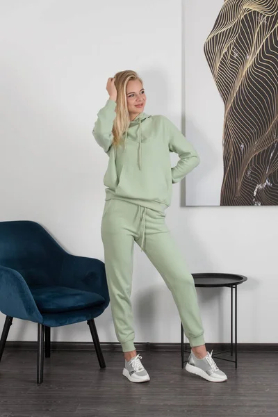 Стильна красива молода блондинка в світло-зеленому костюмі позує біля білої стіни в кімнаті. Приваблива дівчина модель позує біля синього стільця . — стокове фото