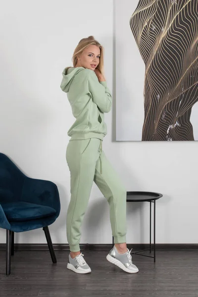 Стильная красивая молодая блондинка в светло-зеленом спортивном костюме позирует возле белой стены в комнате. Привлекательная девушка-модель позирует рядом с синим стулом. — стоковое фото