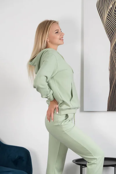 Elegante hermosa mujer rubia joven en un chándal verde claro posa cerca de una pared blanca en la habitación. Modelo chica atractiva posando cerca de la silla azul. —  Fotos de Stock