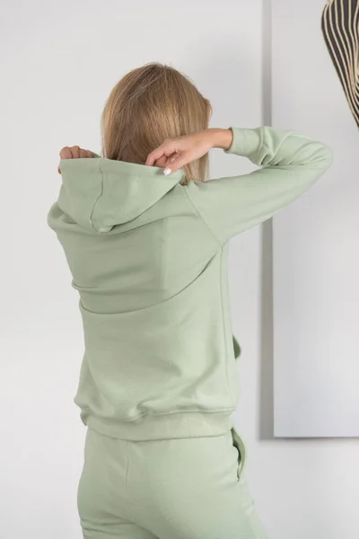 Stijlvolle mooie jonge blonde vrouw in een groen trainingspak poseert in de buurt van een witte muur in de kamer. — Stockfoto