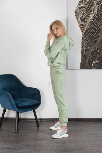 穿着浅绿色运动服的时髦漂亮的金发女人在房间的白墙边摆姿势。迷人的女模特摆出接近蓝色椅子的姿势. — 图库照片