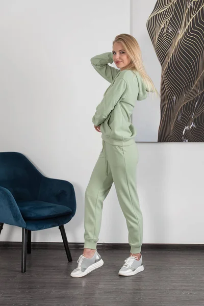 Стильная красивая молодая блондинка в светло-зеленом спортивном костюме позирует возле белой стены в комнате. Привлекательная девушка-модель позирует рядом с синим стулом. — стоковое фото