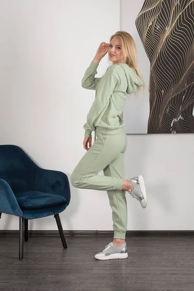 Mulher loira bonita e elegante em um traje verde claro posa perto de uma parede branca na sala. Modelo de menina atraente posando perto da cadeira azul. — Fotografia de Stock