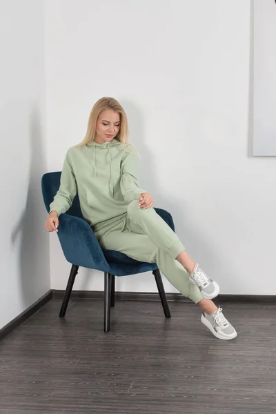 Mulher loira bonita e elegante em um traje verde claro posa perto de uma parede branca na sala. Modelo de menina atraente posando em uma cadeira azul. — Fotografia de Stock