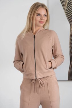 Biege eşofmanlı güzel sarışın kadın odadaki beyaz duvarın yanında poz veriyor. Fitness kadını.