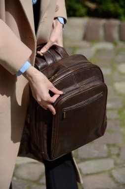 Şık bir kadın model, kahverengi deri bir sırt çantası ve detaylı fermuarlar sergiliyor. Şehir ortamında gelişmiş bej renkli bir trençkotun karşısında duruyor..