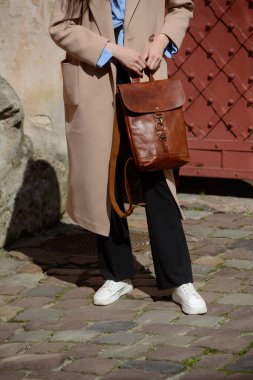 Klasik bej ceketli ve spor ayakkabılı şık bir kadın model, eşsiz bir kapağı ve tokası olan sofistike kahverengi deri bir sırt çantası sergiliyor..