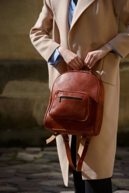 Şık bir kadın model, kahverengi deri bir sırt çantası ve detaylı fermuarlar sergiliyor. Şehir ortamında gelişmiş bej renkli bir trençkotun karşısında duruyor..